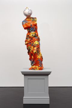 A sculpture on a pedestal dressed in a fabric with an orange pattern in yellows and blues. With a globe as head and no arms.
