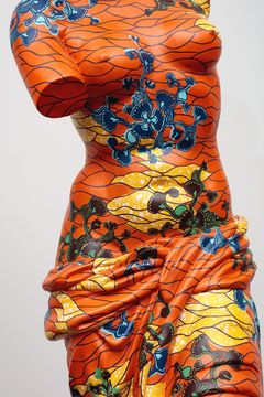 Close up of a sculpture with no arms on a pedestal dressed in a fabric with an orange pattern in yellows and blues.