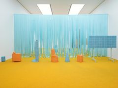 A room with turquoise and orange objects placed on an orange carpet with turquoise strips hanging from the walls in the background.
