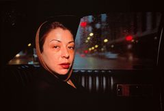 A photography of a woman in the back seat of a taxi. She is looking at the photographer with a serious expression and is dressed in a brown headdress and dark clothes.