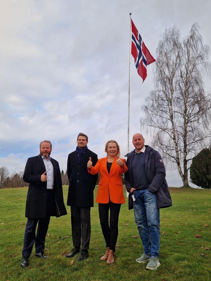 Fotografi av blant annet Kristin Børresen, administrerende direktør i Graminor og Landbruks- og matminister Nils Kristen Sandtrøen foran et heist flagg hos Graminor.