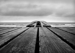 Svart-hvitt-bilde av en person som ligger delvis skjult bak en trebrygge ved havet.