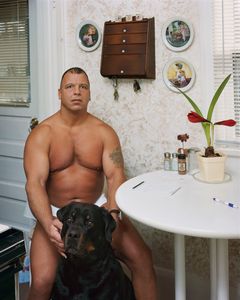 Portrett av en naken mann som sitter med en svart hund ved et lite kjøkkenbord.
