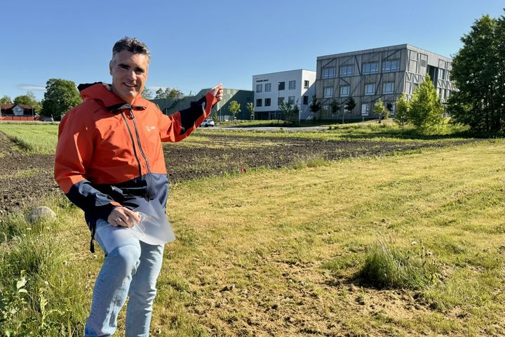 En person i rød jakke står på en gressplen ved siden av et jordbruksområde og peker mot noen bygninger i bakgrunnen.