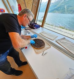 Mann i arbeid ved et vindu fjerner gjærbøs fra en gjærkrans laget av flettet hestehår. Fjordutsikt i bakgrunnen.