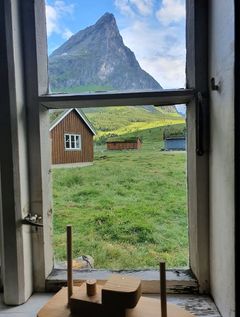 Utsikt fra et vindu over en gresskledd slette med trehus og en bratt fjelltopp i bakgrunnen.