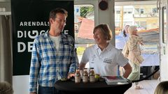 To personer står ved et bord med saftflasker, foran en plakat som sier "Arendals DIALOG Debatt".