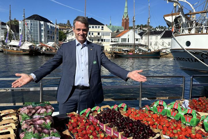 En mann står smilende foran et bord med norske bær ved en havn.