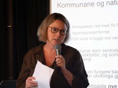 Kvinne holder en mikrofon og papirer mens hun snakker foran en skjerm med tekst på.