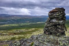 Utsikt over et fjellandskap med variert terreng og en steinvarde i forgrunnen under en dramatisk skyet himmel.