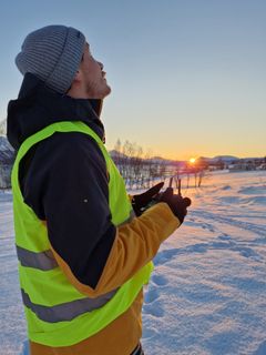 Mann i refleksvest styrer en drone på et snødekt landskap ved solnedgang.
