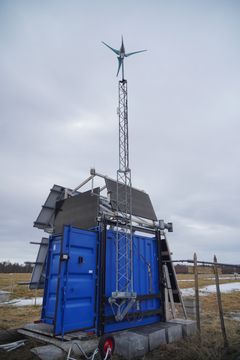 En blå container med solcellepaneler på taket og en vindmølle montert på en mast ved siden av.