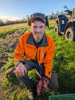 En person i oransje jakke sitter ute i en åker og holder noen søtpoteter.