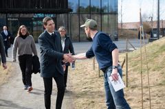 En person håndhilser på en annen person utendørs med flere mennesker i bakgrunnen.