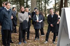 Alt text: En gruppe mennesker står utendørs i en skogkledd område, ved siden av et skilt. De er delaktige i en diskusjon eller presentasjon.