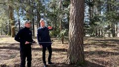 To personer står i en skog, og den ene peker på et tre mens de har en samtale.