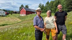 Tre personer står i en blomstereng foran en rød låve og noen hus.