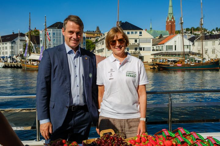 To personer står foran en havn med båter og bygninger i bakgrunnen, ved et bord med frukt og bær.