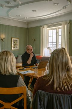 En gruppe mennesker sitter rundt et bord i et rom med grønne vegger. Én person bruker en datamaskin.