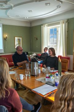 Personer sitter rundt et bord i et konferanserom og snakker sammen. Bordet er dekket med notatbøker, teposer og en vannkaraffel. En projektor er synlig i venstre hjørne.