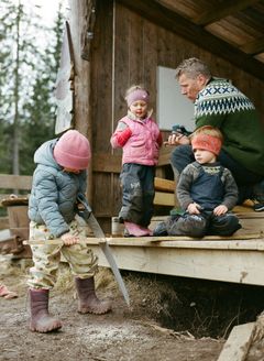 Tre barn og en voksen ved en snekkerbod i skogen. et barn i rosa lue og blå dunjakke sager i en planke med en stor håndsag, et barn hjelper ved å sitte på planken, et barn står på plattingen og ser på, mens en voksen mann i grønn strikkegenser viser noe til det ene barnet.