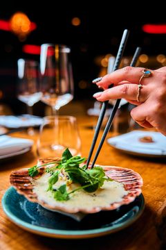 En hånd bruker spisepinner for å spise grillede kamskjell med grønnsaker, servert på en tallerken.