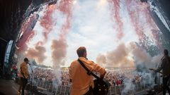 En konsertscene med musikere som spiller for et stort publikum, omgitt av røykeffekter.