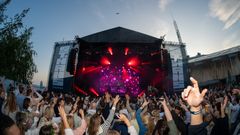 Konsert med publikum som strekker hendene opp, rød scenebelysning og fargerike lysstråler ved solnedgang.