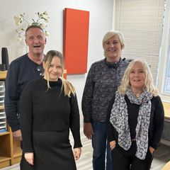 Fire personer som står innendørs foran et vindu og et bord.