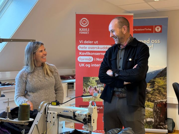 To personer smiler mens de står ved en symaskin. Bak dem er bannere med informasjon om en stiftelse.