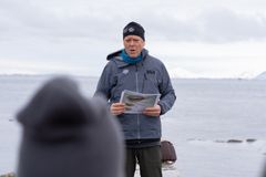 En person står utendørs ved sjøen, snakker og holder papirer.