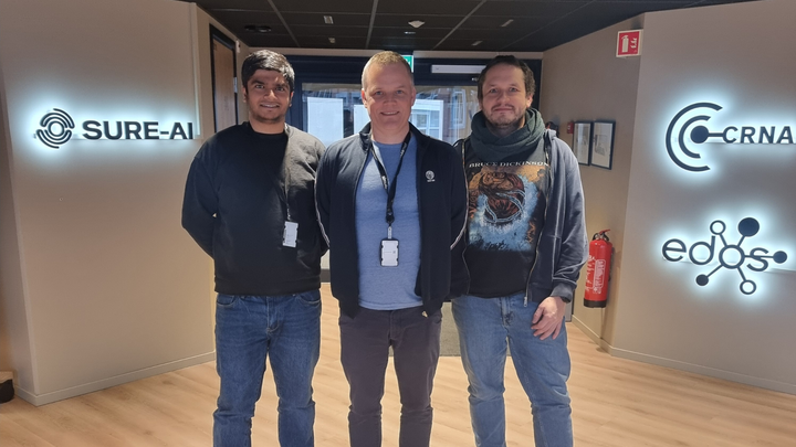 Forskerne Sushant Gautam, Klas Pettersen og Michael Riegler står ved inngangen til kontorlokalet for SimulaMet.