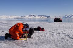 En person i en oransje drakt utfører feltarbeid på isen i Kongsfjorden, Svalbard, med fjell og en båt i bakgrunnen.