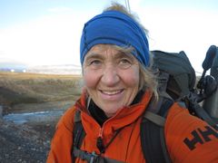 En person iført oransje jakke, blått pannebånd og ryggsekk smiler utendørs i et arktisk landskap.