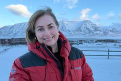 En smilende kvinne i rød jakke med snødekte fjell i bakgrunnen.