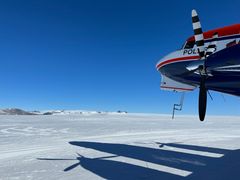 Et fly står på en isdekket flate under en klar blå himmel. Flyets skygge vises på isen. Fjell kan sees i bakgrunnen.