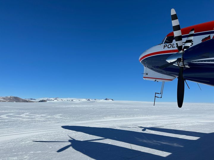 Et fly står på en isdekket flate under en klar blå himmel. Flyets skygge vises på isen. Fjell kan sees i bakgrunnen.