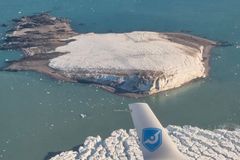 Et flyvindu viser en isdekket øy i et turkis hav, med isflak spredt rundt.