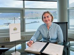 Person sitter ved et skrivebord med dokumenter og en penn, med utsikt over en havn i bakgrunnen.