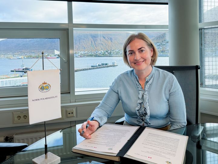 Person sitter ved et skrivebord med dokumenter og en penn, med utsikt over en havn i bakgrunnen.