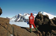 Person i rødt yttertøy på steinete terreng foran snødekte fjell og isbre.