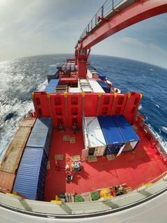 Fiskeøyefoto av dekket på et rødt containerskip med last og containere, sett fra masten, med havet rundt.