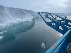 Alt-tekst: En blå kran fra et forskningsskip senker et måleinstrument ned i havet ved foten av en høy, hvit isbrem i Antarktis.