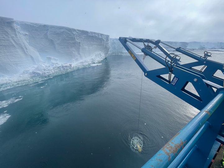 Alt-tekst: En blå kran fra et forskningsskip senker et måleinstrument ned i havet ved foten av en høy, hvit isbrem i Antarktis.
