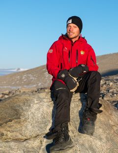 Mann i rød polarjakke og lue sitter på en stor stein i et polart landskap med snø og fjell i bakgrunnen.