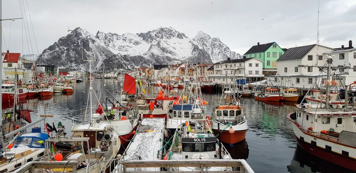 Bilde av en snødekt fiskerihavn i Henningsvær, omgitt av fiskebåter og hus med fjell i bakgrunnen.