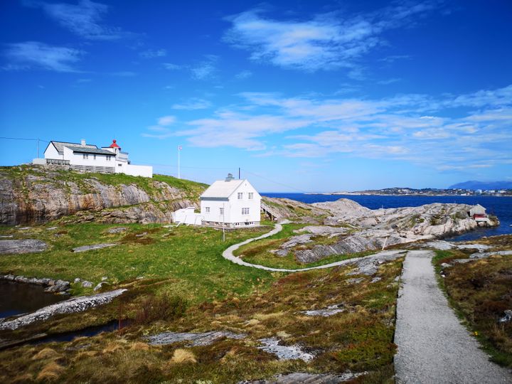 To hvite hus ved en steinete kystlinje under en blå himmel. En sti fører mot husene, og havet er synlig i bakgrunnen.
