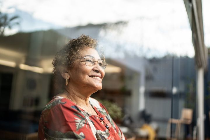 Eldre dame ser smilende ut gjennom vinduet fra boligen sin. Foto.