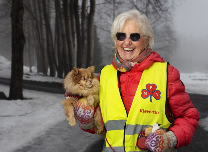 Eldre, smilende kvinne med Sanitetskvinnenes refleksvest går tur i tåka, og bærer på en liten hund