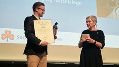 Mann og kvinne på scene. Mann holder diplom og statuett.